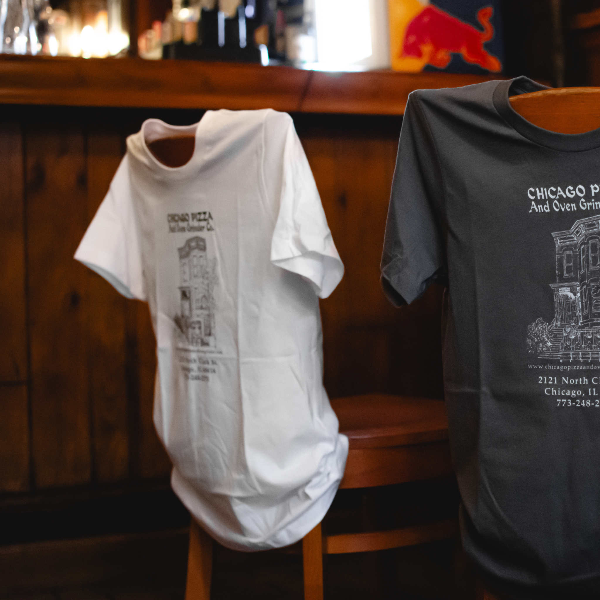 Two t-shirts, one white and one gray, with printed designs on a wooden stool against a bar background.