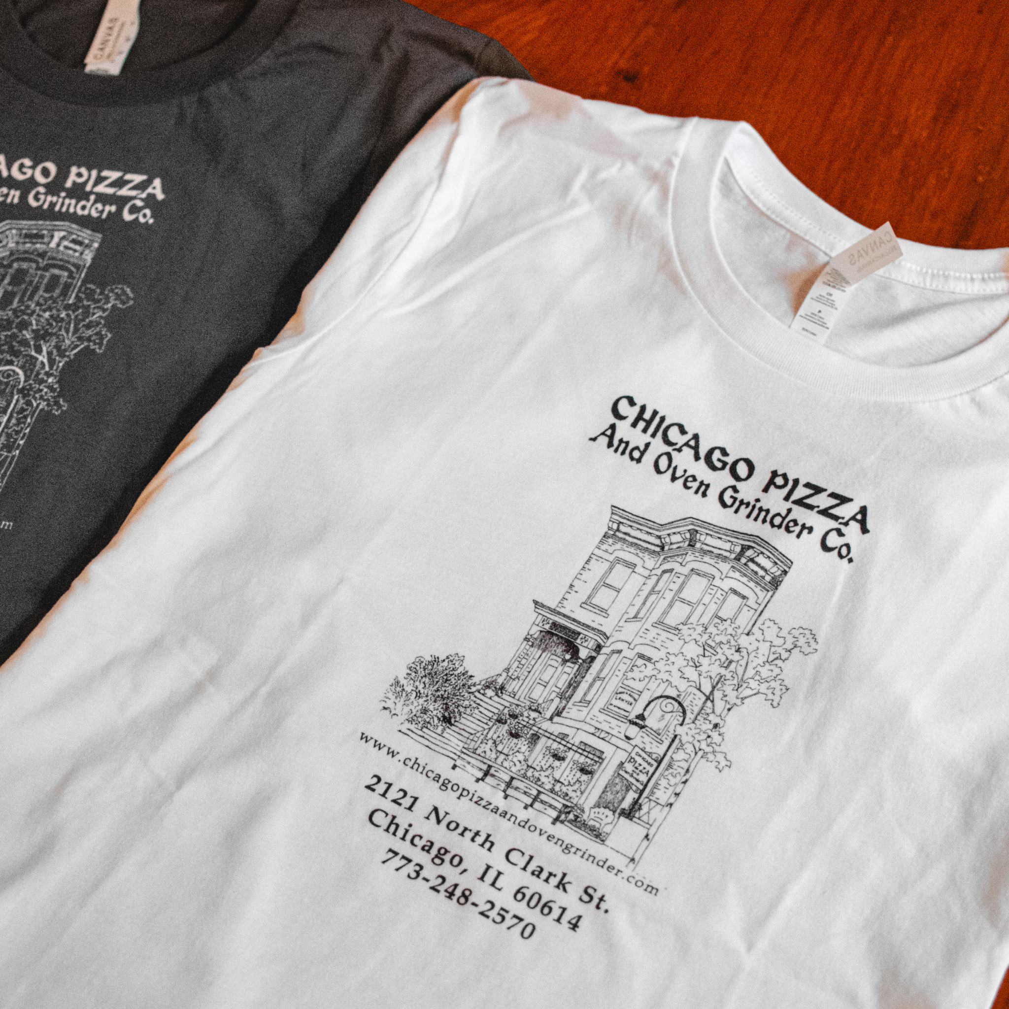 Two t-shirts with 'Chicago Pizza and Oven Grinder Co.' design on a wooden surface